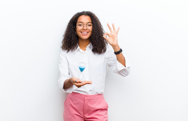 Joven mujer bastante negra con un reloj de arena contra la pared blanca