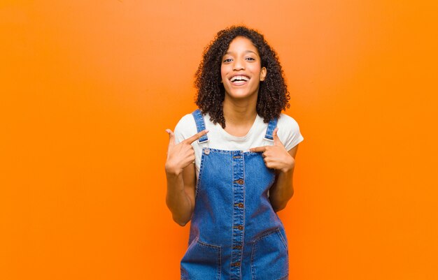 Joven mujer bastante negra sintiéndose feliz, sorprendida y orgullosa, apuntando a sí misma con una mirada emocionada y asombrada contra la pared naranja