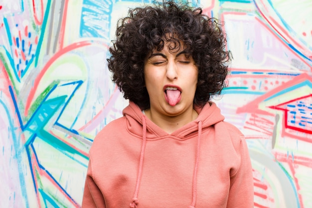 Joven mujer bonita afro con actitud alegre, despreocupada, rebelde, bromeando y sacando la lengua, divirtiéndose contra el graffiti