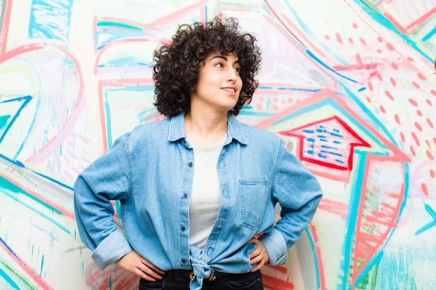 Joven mujer bonita afro mirando feliz alegre y confiada sonriendo con orgullo y mirando al lado con ambas manos en las caderas contra la pared de graffiti