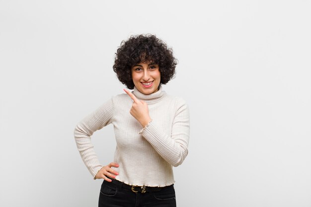 Joven mujer bonita afro sonriendo alegremente, sintiéndose feliz y apuntando hacia un lado y hacia arriba, mostrando el objeto en el espacio de la copia