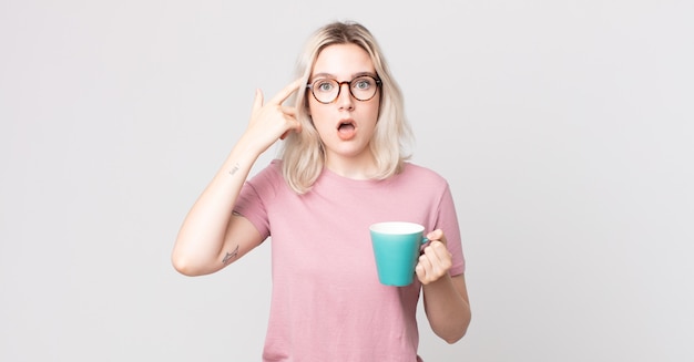 Joven mujer bonita albina mirando sorprendido, dándose cuenta de un nuevo pensamiento, idea o concepto con una taza de café