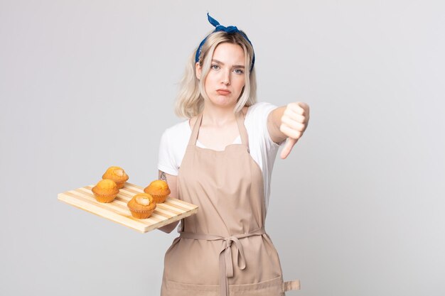 Joven mujer bonita albina que se siente cruzada, mostrando los pulgares hacia abajo con una bandeja de muffins