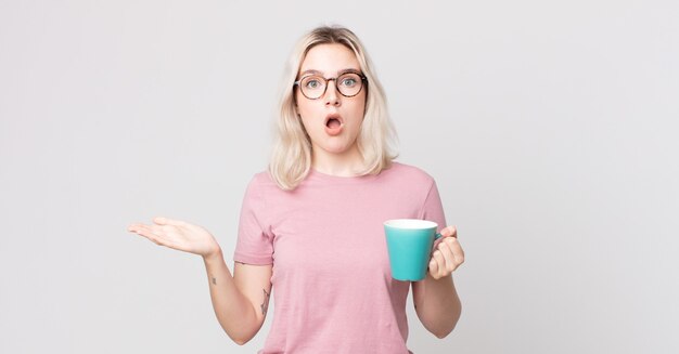 Joven mujer bonita albina que se siente extremadamente conmocionada y sorprendida con una taza de café