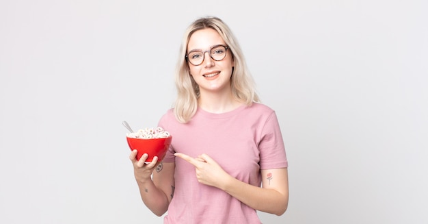 Joven mujer bonita albina sonriendo alegremente, sintiéndose feliz y apuntando hacia un lado con un tazón de desayuno