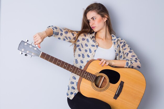 Joven mujer bonita con blusa beige y pantalones negros arreglando una guitarra con fondo gris en el estudio