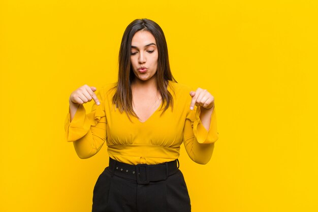 Joven mujer bonita con la boca abierta apuntando hacia abajo con ambas manos, mirando sorprendido, asombrado y sorprendido aislado contra la pared naranja