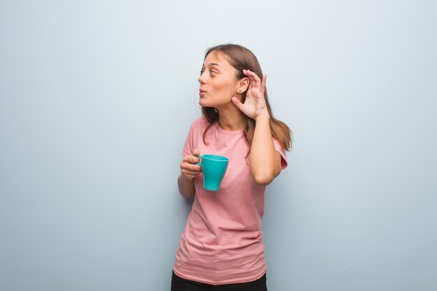 Joven mujer bonita caucásica intenta escuchar un chisme. Ella sostiene una taza.