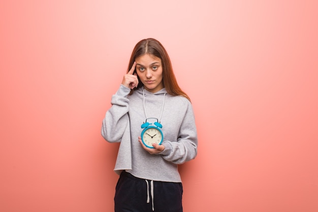 Joven mujer bonita caucásica pensando en una idea. Ella sostiene un despertador.