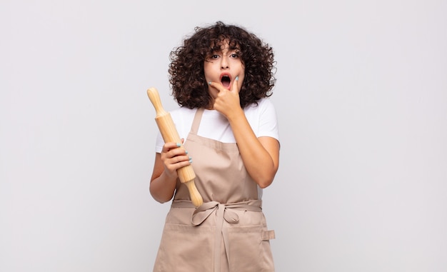 Joven mujer bonita chef con la boca y los ojos bien abiertos y la mano en la barbilla, sintiéndose desagradablemente conmocionado, diciendo qué o guau