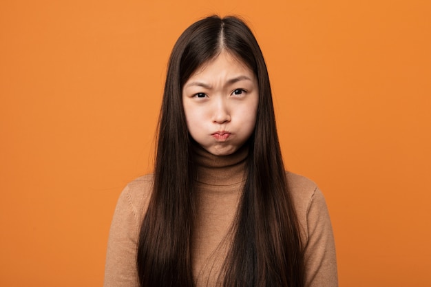 Joven mujer bonita china sopla mejillas, tiene expresión cansada.