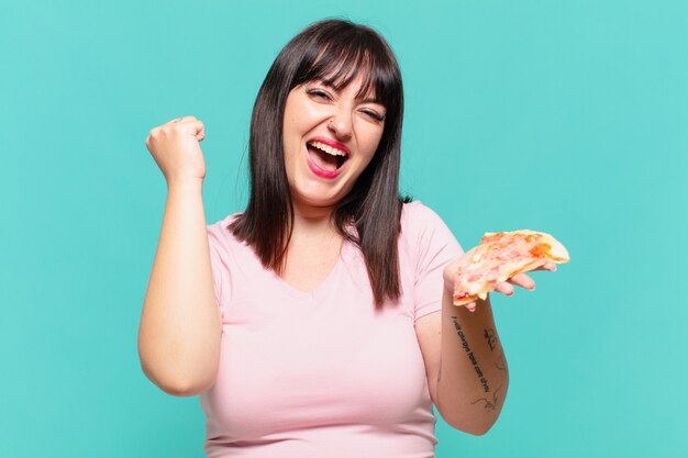 Joven mujer bonita con curvas celebrando una victoria exitosa y sosteniendo una pizza