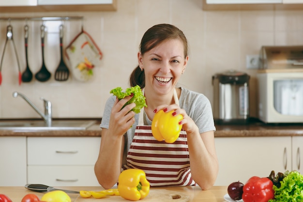 La joven mujer bonita en un delantal con hojas de lechuga y pimiento amarillo riendo en la cocina. Concepto de dieta. Estilo de vida saludable. Cocinar en casa. Prepara comida.