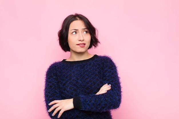 Joven mujer bonita dudando o pensando, mordiéndose el labio y sintiéndose insegura y nerviosa, buscando copiar el espacio en el costado contra la pared rosa