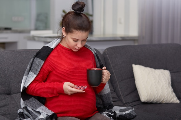 Joven mujer bonita embarazada toma medicamentos, sostiene la taza con bebida, necesita vitaminas, usa ropa casual, posa en el sofá