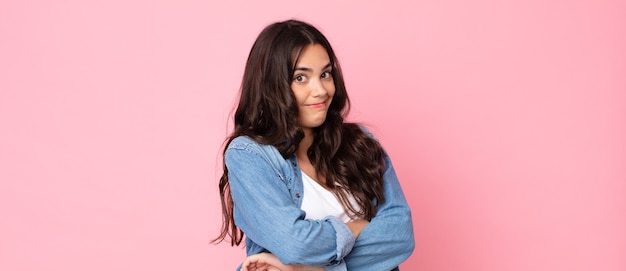 Joven mujer bonita encogiéndose de hombros, sintiéndose confundida e insegura, dudando con los brazos cruzados y la mirada perpleja