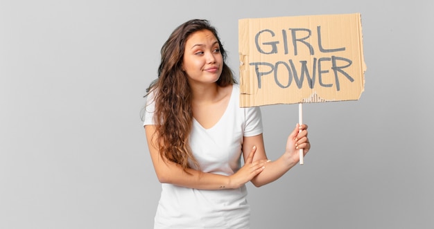 Foto joven mujer bonita encogiéndose de hombros, sintiéndose confundida e insegura y sosteniendo una pancarta de poder femenino