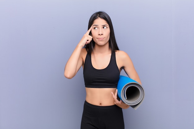 Joven mujer bonita con una estera de yoga contra la pared de cemento