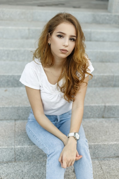 Joven mujer bonita con estilo en jeans vintage de moda en una camiseta blanca con estilo está descansando sentado en los escalones