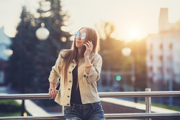 Joven mujer bonita con estilo manos sosteniendo un teléfono Fondo de puesta de sol Día soleadobuenas gafas de sol accesorios geniales