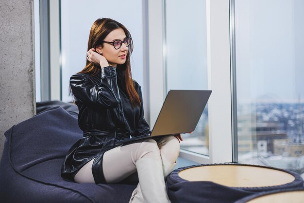 Joven mujer bonita con gafas está trabajando en una computadora portátil mientras está sentada en un espacio de trabajo moderno Trabajo remoto independiente