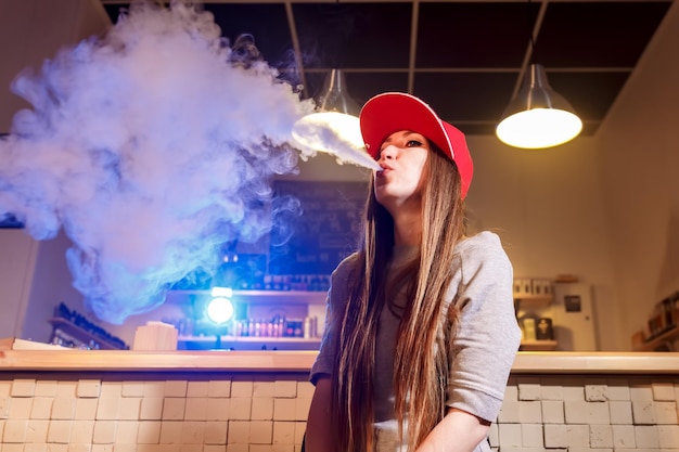 Joven mujer bonita con gorra roja fumar un cigarrillo electrónico en la tienda de vape