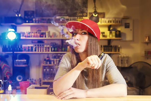 Joven mujer bonita con gorra roja fumar un cigarrillo electrónico en la tienda de vape