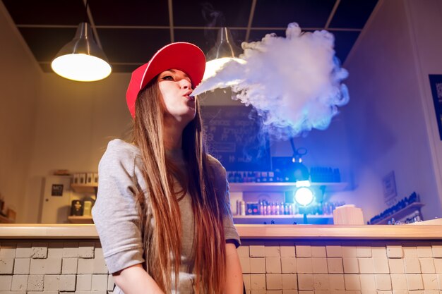 Joven mujer bonita con gorra roja fumar un cigarrillo electrónico en la tienda de vape