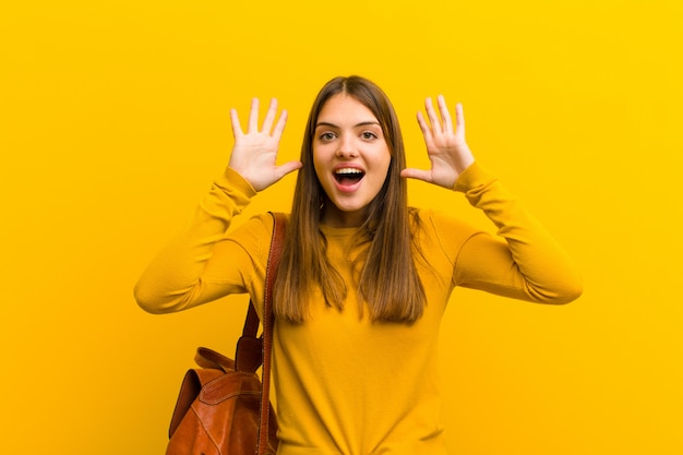 Joven mujer bonita gritando de pánico o ira, sorprendida, aterrorizada o furiosa, con las manos al lado de la cabeza contra la pared naranja