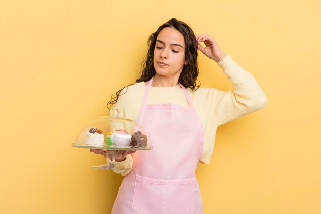 Joven mujer bonita hispana que se siente perpleja y confundida rascando el concepto de jefe de cocina y pasteles