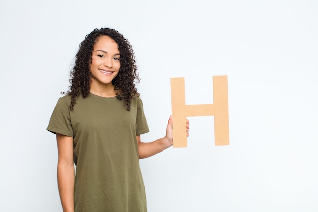 Joven mujer bonita latina emocionada, feliz, alegre, sosteniendo la letra H del alfabeto para formar una palabra o una oración