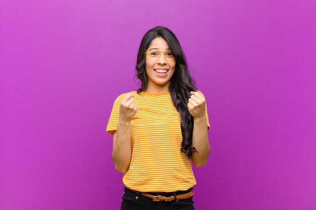Joven mujer bonita latina gritando triunfante, riendo y sintiéndose feliz y emocionada mientras celebra el éxito contra la pared púrpura