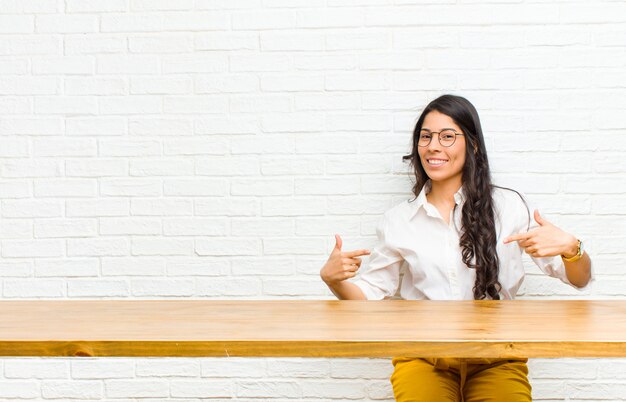 Joven mujer bonita latina que parece orgullosa, arrogante, feliz, sorprendida y satisfecha, apuntando a sí misma, sintiéndose como una ganadora sentada frente a una mesa