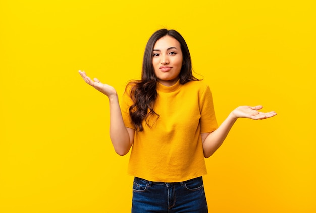 Joven mujer bonita latina que parece perpleja, confundida y estresada, preguntándose entre diferentes opciones, sintiéndose insegura contra la pared plana