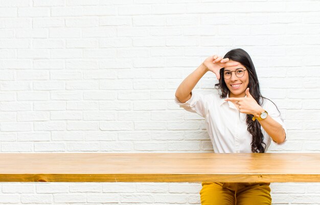 Foto joven mujer bonita latina se siente feliz, amigable y positiva, sonriendo y haciendo un marco de foto de retrato con las manos sentado frente a una mesa