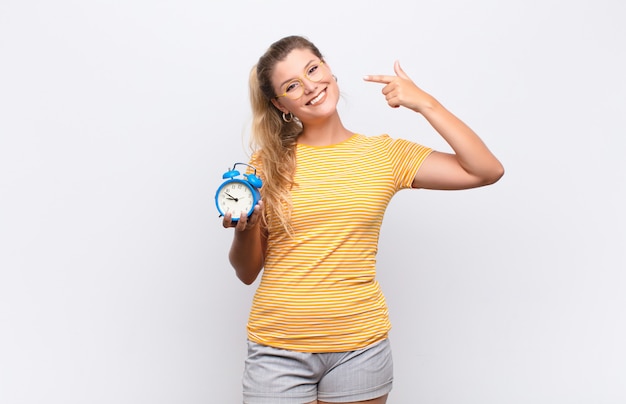 Joven mujer bonita latina sonriendo con confianza apuntando a su propia sonrisa amplia, actitud positiva, relajada y satisfecha con un reloj despertador