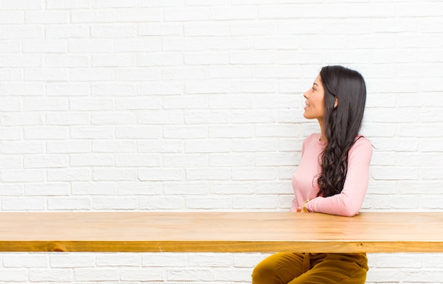 Joven mujer bonita latina en vista de perfil buscando copiar espacio por delante, pensando, imaginando o soñando despierto sentado frente a una mesa