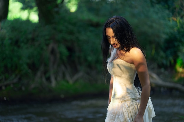 Joven mujer bonita morena en vestido de novia blanco al aire libre cerca del río con pelos mojados