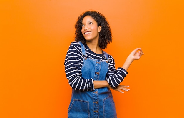 Joven mujer bonita negra sonriendo alegremente y mirando hacia los lados, preguntándose, pensando o teniendo una idea contra la pared naranja