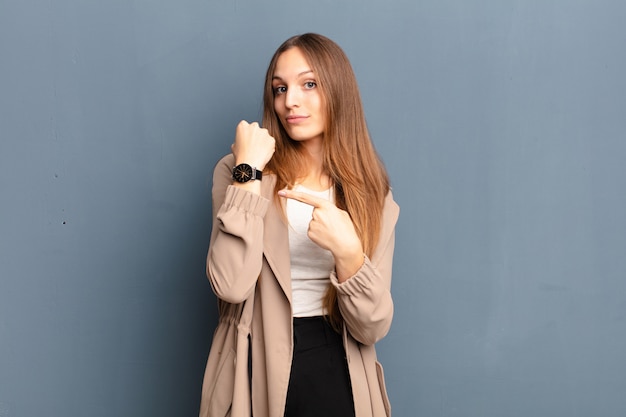 Joven mujer bonita que parece impaciente y enojada, apuntando al reloj, pidiendo puntualidad, quiere llegar a tiempo sobre la pared gris