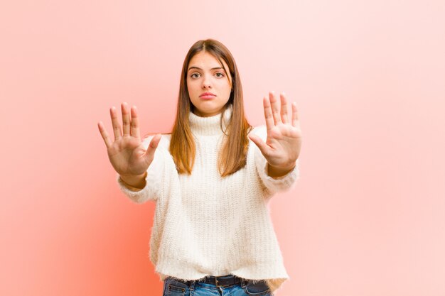 Joven mujer bonita que parece seria, infeliz, enojada y disgustada prohibiendo la entrada o diciendo que pare con ambas palmas abiertas contra la pared rosa