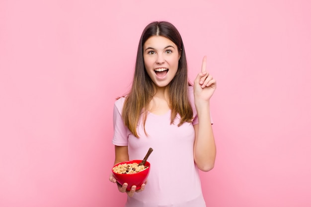 Joven mujer bonita que se siente como un genio feliz y emocionado después de darse cuenta de una idea, levantando alegremente el dedo, ¡eureka! con un tazón de desayuno