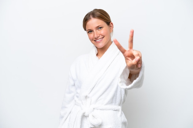 Foto joven mujer bonita rubia en bata de baño aislado sobre fondo blanco sonriendo y mostrando el signo de la victoria