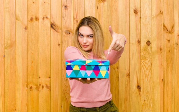 Joven mujer bonita rubia con una caja de regalo contra la pared de madera