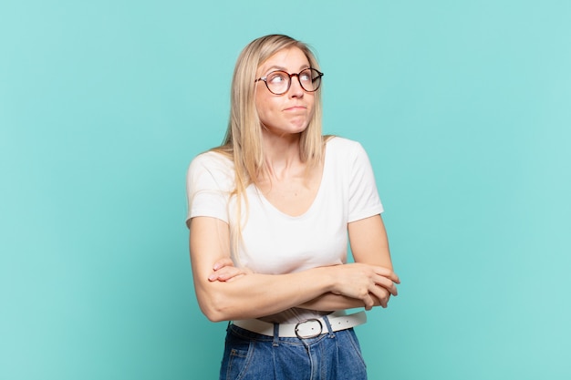 Joven mujer bonita rubia encogiéndose de hombros, sintiéndose confundida e insegura, dudando con los brazos cruzados y la mirada perpleja