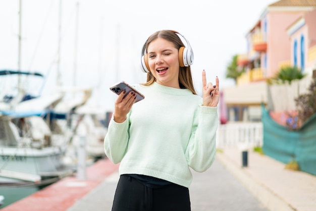 Joven mujer bonita rubia escuchando música con un móvil haciendo gesto de rock