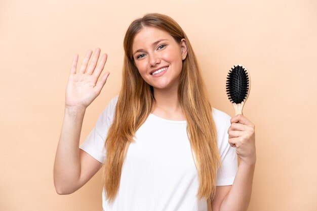 Joven mujer bonita rubia con peine aislado sobre fondo beige saludando con la mano con expresión feliz