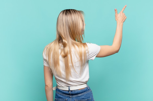 Foto joven mujer bonita rubia de pie y apuntando al objeto en el espacio de la copia, vista trasera