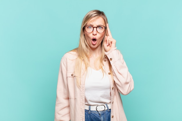 Joven mujer bonita rubia que parece sorprendida, con la boca abierta, conmocionada, dándose cuenta de un nuevo pensamiento, idea o concepto