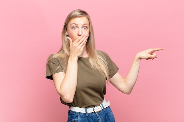 Joven mujer bonita rubia que se siente feliz, conmocionada y sorprendida, cubriendo la boca con la mano y apuntando al espacio lateral de la copia